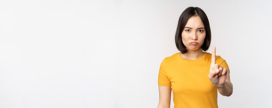 Portrait of asian woman looking serious and angry, showing stop prohibit gesture, taboo sign, forbidding smth, standing in yellow tshirt over white background.