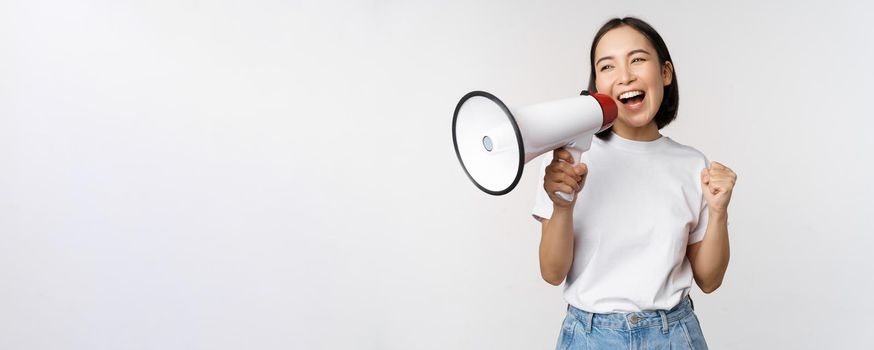 Asian girl shouting at megaphone, young activist protesting, using loud speakerphone, making announcement, white background. Copy space