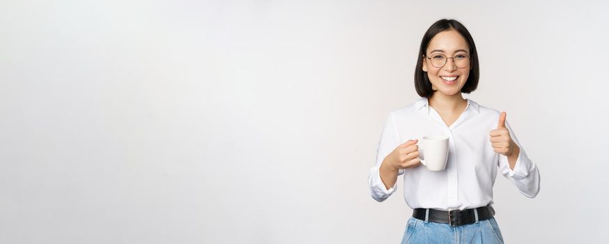 Smiling asian girl holding mug, white cup and thumbs up, recommending drink, coffee or tea, standing over white background.