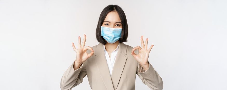 Coronavirus and workplace concept. Image of asian saleswoman, company worker in medical mask showing okay sign, smiling pleased, white background.