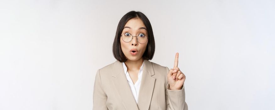 Excited asian woman in glasses, raising finger, eureka sign, has an idea, standing over white background. Copy space
