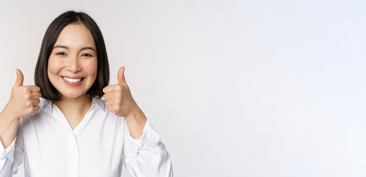Cheerful asian woman face, showing thumbs up and smiling, pleased by something, recommending smth good, standing over white background.