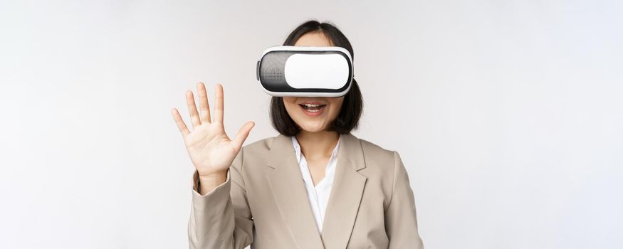 Meeting in vr chat. Asian businesswoman in virtual reality glasses, waving hand and saying hello, greeting someone, standing over white background.