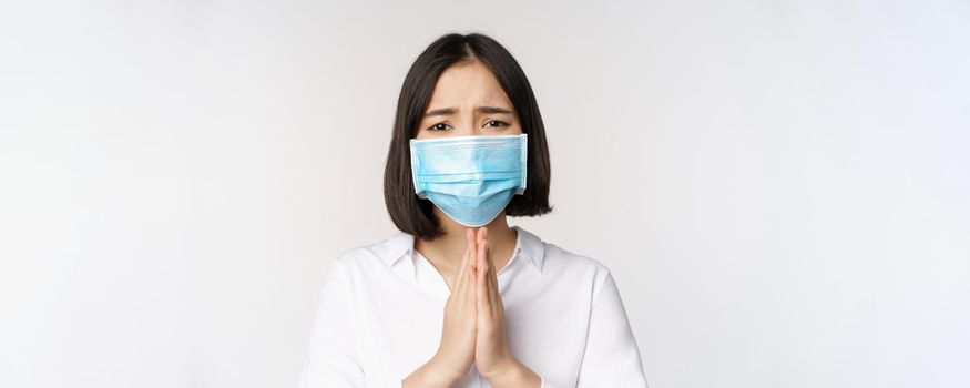 Portrait of asian woman in medical face mask from covid, begging, asking for help, say please, standing over white background.