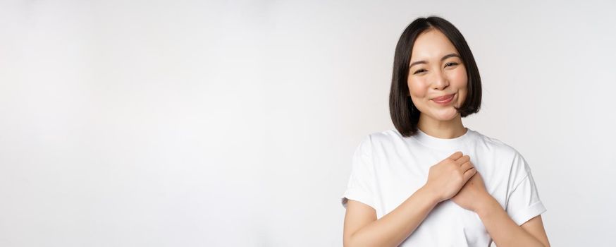 Portrait of carig young asian woman holding hands on heart, gazing with care and love, touched by something, feel flattered, standing over white background.