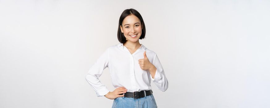 Image of confident asian woman showing thumb up in approval, recommending, like smth good, standing over white background.