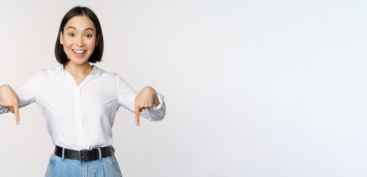 Portrait of young asian woman pointing fingers down and smiling, showing banner, click on link below gesture, inviting people to follow, standing over white background.