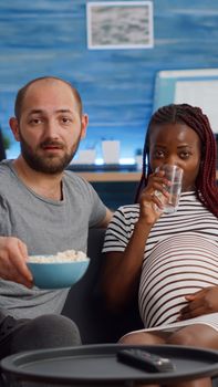 POV of interracial family looking at camera sitting on couch. Mixed race couple expecting child watching television. Caucasian man bringing glass of water for black pregnant woman