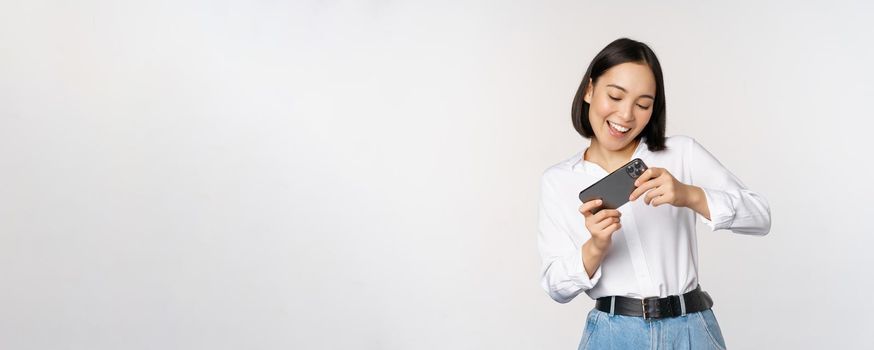 Young korean woman, asian girl playing mobile video game on smartphone, looking at horizontal phone screen, standing over white background.