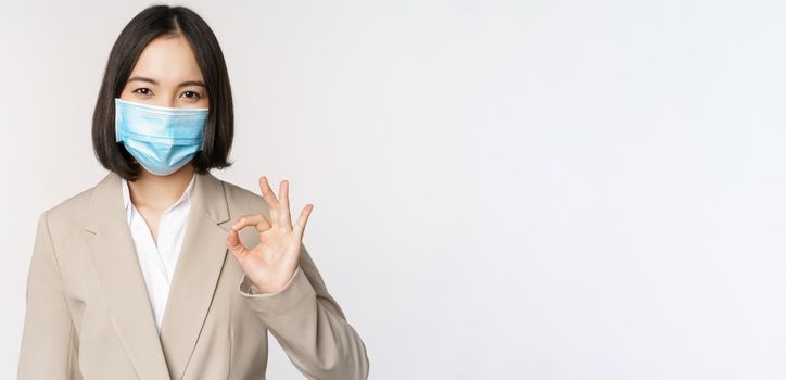 Coronavirus and workplace concept. Image of asian saleswoman, company worker in medical mask showing okay sign, smiling pleased, white background.