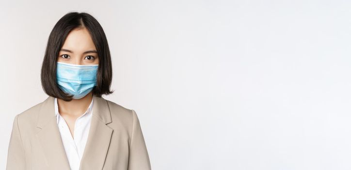 Coronavirus and workplace concept. Portrait of asian office worker, businesswoman in medical face mask looking at camera, white background.