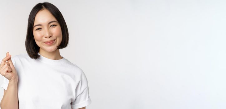 Beautiful asian woman smiling, showing finger hearts gesture, wearing tshirt, standing against white background.