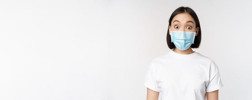 Health and covid pandemic concept. Image of korean woman in face medical mask looking surprised, reacting amazed, standing over white background.
