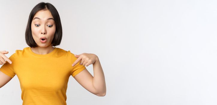 Happy asian girl pointing fingers down, looking at announcement, banner or advertisment, standing in yellow tshirt over white background.
