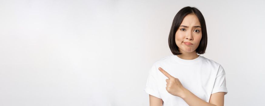 Skeptical asian girl in white t-shirt, pointing at product or logo with disappointed grimace, dislike and complain at smth, standing over white background.