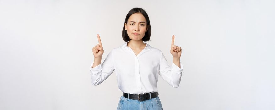 Image of skeptical asian lady grimacing, frowning upset, pointing fingers up, showing advertisement, standing over white background.