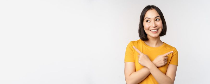 Cute asian girl pointing fingers sideways, showing left and right promo, two choices, variants of products, standing in yellow tshirt over white background.