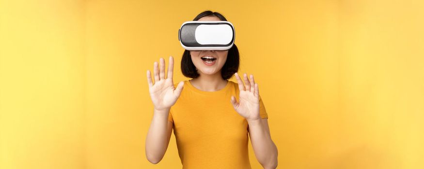 Young asian woman playing in VR, using virtual reality glasses, wearing yellow t-shirt against studio background. Copy space