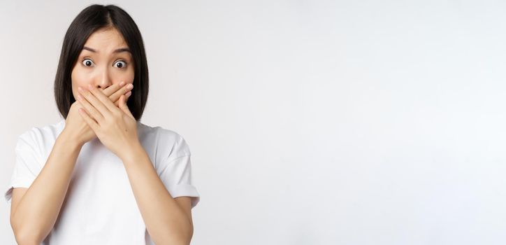 Image of shocked young asian woman shut mouth, looking startled, speechless, standing over white background. Copy space
