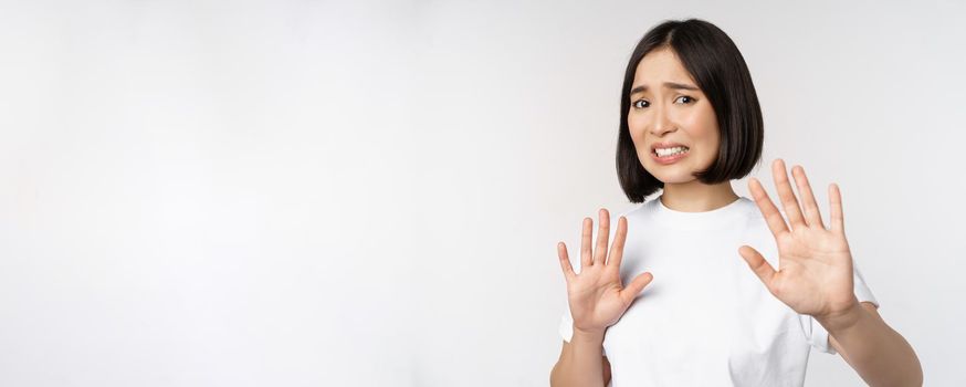 Disgusted asian woman rejecting smth, grimacing from dislike and aversion, stare with cringe, refusing offer, standing over white background.