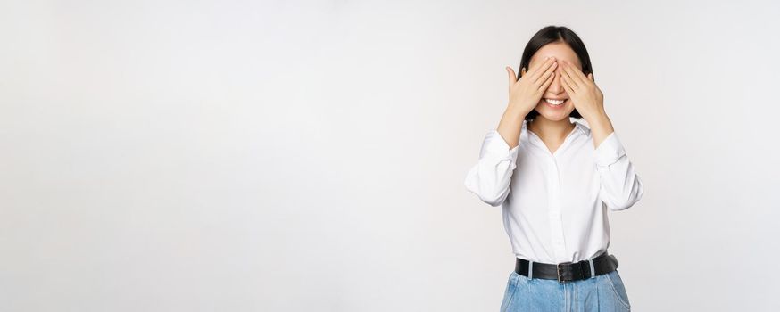 Image of young asian woman close her eyes and smiles, waits for surprise, anticipates, stands over white background.