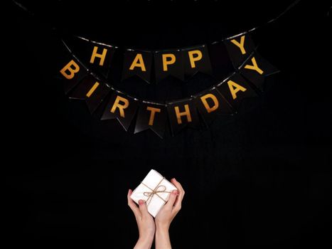 Faceless person holding a white gift in his hands on a black isolated background with the inscription in gold letters Happy Birthday. This photo shows a universal birthday greeting. High quality photo