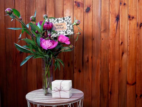 Pink flowers peonies in a vase on a wooden background with a small white gift and a happy birthday card. The photo was taken with natural daylight.