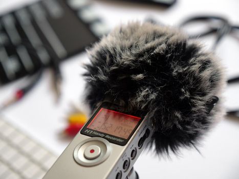 Record audio inside on a zoom h1 recorder with a dead cat in the studio in natural daylight. The background shows items such as Clapperboard, keyboard, cord, cable, 3.5, mini jack. You can use this photo in any project related to sound recording, podcast, recording studio, interview