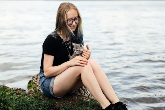 Young blonde woman with blue hair, wearing glasses, denim shorts and a black T-shirt. Sitting on a floating tree near the lake with a three-month-old kitten breed Scottish Straight.