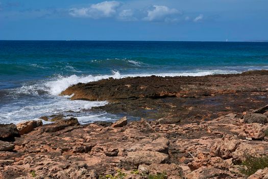 Rocks on the shore hit by the waves. Balearic Islands, Mediterranean Sea, blue sky, splashes of water.