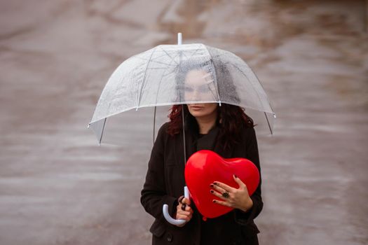 Sad middle-aged woman in a black coat and boots under a transparent umbrella. He holds a balloon in the form of a heart in his hands. Rain in the city, outdoor scene, water drops on the umbrella. The concept of loneliness, unhappy love