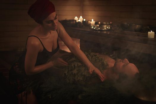 A woman conducts a bathing ritual for a man with the help of brooms. A man lies on spruce branches.