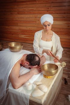 A woman performs Tibetan singing bowl therapy with a man lying under a white sheet. Relaxing meditation.