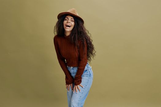 Enjoyed stylish curly Latin female in brown hat, smiling at camera, holding hand on hips, laughing, saying Yes, isolated green background. Copy space clothing fashion brands, free place for your ad.
