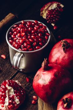 Rustic metal mug full of pomegranate seeds. Whole fruits and pomegranate pieces on dark wooden table.