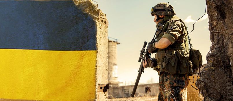 Ukrainian Soldier military in the war with a weapon in his hands. The flag of Ukraine is painted on a brick wall. Relations between Ukraine and Russia.