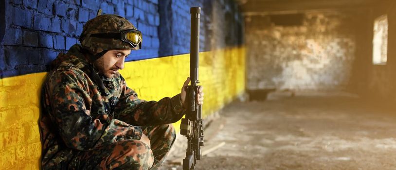 Military soldier on the background of the flag of Ukraine. The flag of Ukraine is painted on a brick wall, a tired sad soldier sits with a weapon in his hands. Relations between Ukraine and Russia.