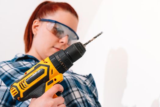 young red-haired girl with her hair tied back, low angle, wearing a blue checked shirt and safety glasses, standing, holding a yellow drill in her right hand to hang a shelf at home on a white wall.