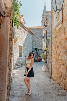 Girl tourist walking through ancient narrow street on a beautiful summer day in MEDITERRANEAN MEDIEVAL CITY , OLD TOWN bUDVA, MONTENEGRO. Young beautiful cheerful woman walking on old street at tropical town. Pretty girl looking at you and smiling