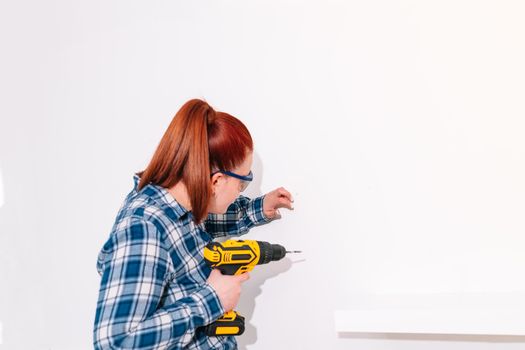 young red-haired woman with her hair up, flat with her back to the camera, wearing blue checked shirt and safety glasses, standing, holding a yellow drill in her right hand to hang a shelf at home on a white wall.