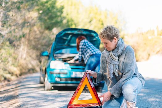 Mother and daughter next to the broken down car, while the blonde adult woman puts the safety triangle in the foreground, the red-haired young woman tries to repair the car with the bonnet open in the background. Two friends on holiday have a car accident and have to call the mechanic in the middle of the road. Clear road on a very sunny day next to a forest with a mountain in the background.