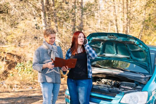 Mother and daughter standing next to the broken down vehicle checking the documents and calling the tow truck. Two friends on holiday have a car accident and have to call the mechanic in the middle of the road. Clear road on a very sunny day next to a forest in the background. Red-haired girl calling tow truck and blonde woman looking at insurance papers.