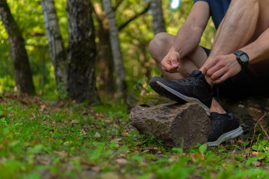 Man tying his shoelaces in the park outdoors, around the forest, oak trees green grass young enduring athletic athlete. active sport forest, lifestyle trail endurance legs marathon, woods Adult energy runners stretches