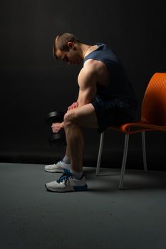 Man on black background sits on a chair rocking his biceps keeps dumbbells pumped up in fitness bodybuilding black, body man athletic heavy, pectoral. Young sportive metal, one fit