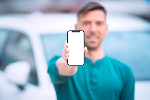 Smartphone mockup, blank screen. Young smiling man standing near white car outdoors and showing or holding his phone with blank space. High quality photo