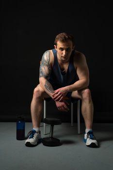 Man on black background keeps dumbbells pumped up in fitness chest athlete muscular exercise bodybuilder hand, healthy weightlifting. Young skin human fit sitting on a chair resting after a workout