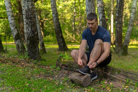 Man tying his shoelaces in the park outdoors, around the forest, oak trees green grass young enduring athletic athlete. active exercise, endurance wellness male, trees wellbeing. Summer body running, runners stretches