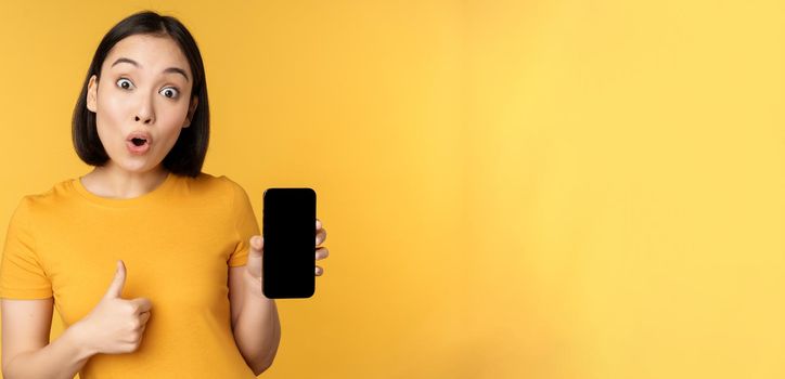 Excited asian woman showing mobile phone screen, thumbs up, like smth good, recommending smartphone application, standing over yellow background.