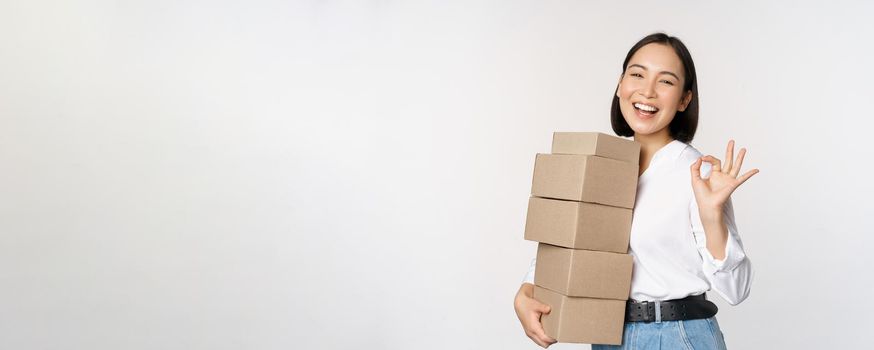 Portrait of beautiful happy woman carry boxes with orders. Businesswoman with carry delivery and showing okay sign, white background.