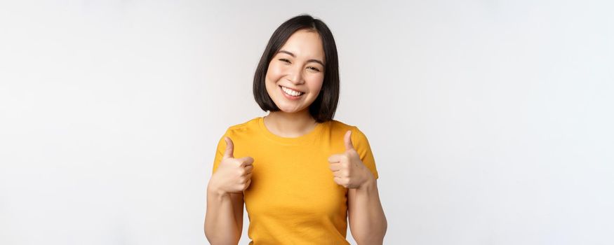 Beautiful smiling asian female model, showing thumbs up and looking pleased, recommending, express positive feedback, standing over white background.